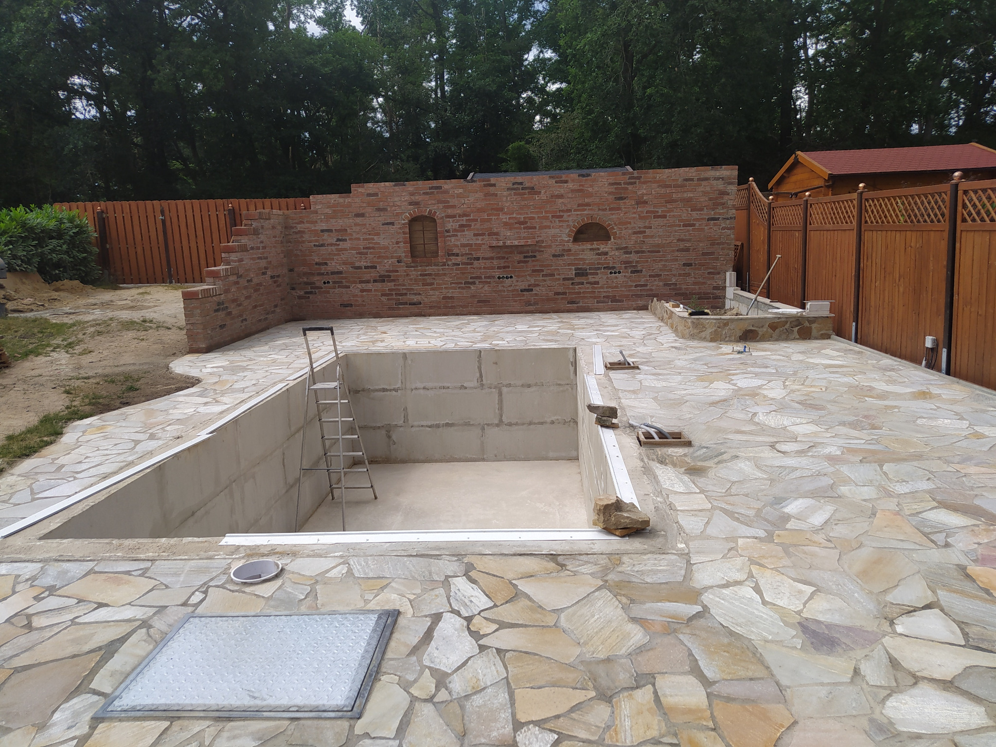 Budowa betonowego basenu z wykończeniem z kamienia naturalnego, widoczna metalowa drabinka, ceglana ściana z dwoma okienkami w tle oraz drewniany płot.