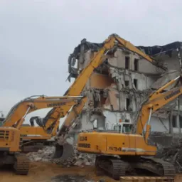 Dwa żółte koparki Liebherr 934 na gąsienicach pracują przy częściowo zburzonym, wielopiętrowym budynku biurowym, odsłaniając jego konstrukcję; w tle pochmurne niebo.