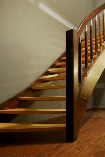 Wolnostojące schody z jasnego drewna z ciemnobrązową ramą i metalowymi słupkami balustrady, oświetlone światłem padającym z góry.