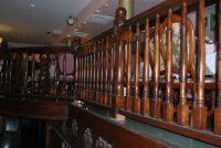 Klasyczna, lakierowana barierka z ciemnego drewna z ozdobnymi tralkami, zabezpieczająca antresolę w pomieszczeniu z ciemnym oświetleniem.