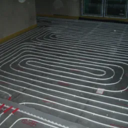 Białe rury ogrzewania podłogowego ułożone spiralnie na szarej izolacji z czerwonymi znacznikami, widok z góry w surowym wnętrzu.