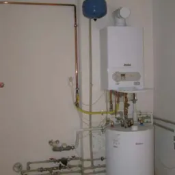 Biały piec gazowy Vaillant z podłączonymi rurami miedzianymi i plastikowymi, niebieski zbiornik wyrównawczy, instalacja w pomieszczeniu z białą ścianą i drzwiami.