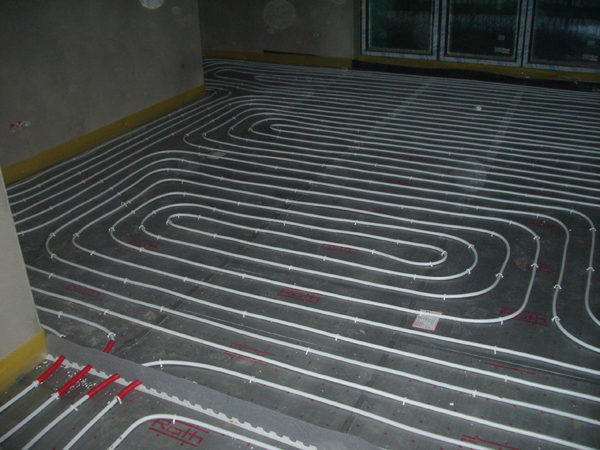 Białe rury ogrzewania podłogowego ułożone spiralnie na szarej izolacji z czerwonymi znacznikami, widok z góry w surowym wnętrzu.