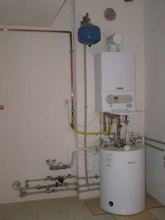 Biały piec gazowy Vaillant z podłączonymi rurami miedzianymi i plastikowymi, niebieski zbiornik wyrównawczy, instalacja w pomieszczeniu z białą ścianą i drzwiami.