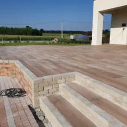 Rozległy taras wyłożony płytami imitującymi drewno, z niskim murkiem z cegły i schodami z szarej kostki, widok na zielony ogród i dom z białymi kolumnami w słoneczny dzień.