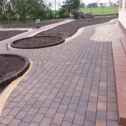 Brązowo-szara kostka brukowa ułożona w łuki tworzące ścieżkę i obramowanie rabat z ziemią, jasna kostka oddziela rabaty od trawnika, brązowe schody prowadzą do budynku.