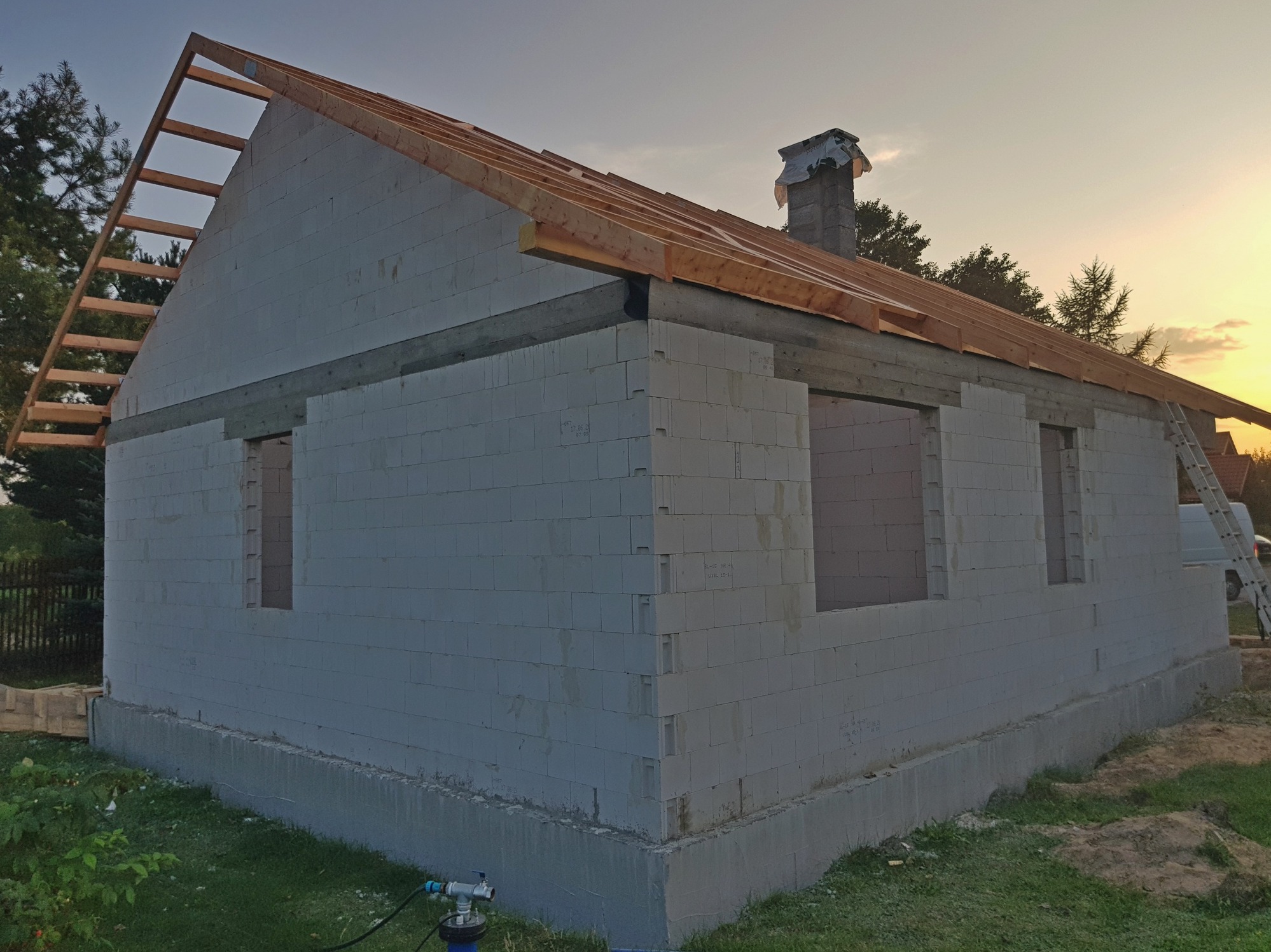 Surowy stan domu z betonu komórkowego w trakcie budowy, widoczne niezadaszone okna, komin z zabezpieczeniem, częściowo zamontowana więźba dachowa, drabina oparta o ścianę, trawa i drzewa w tle, wąż...