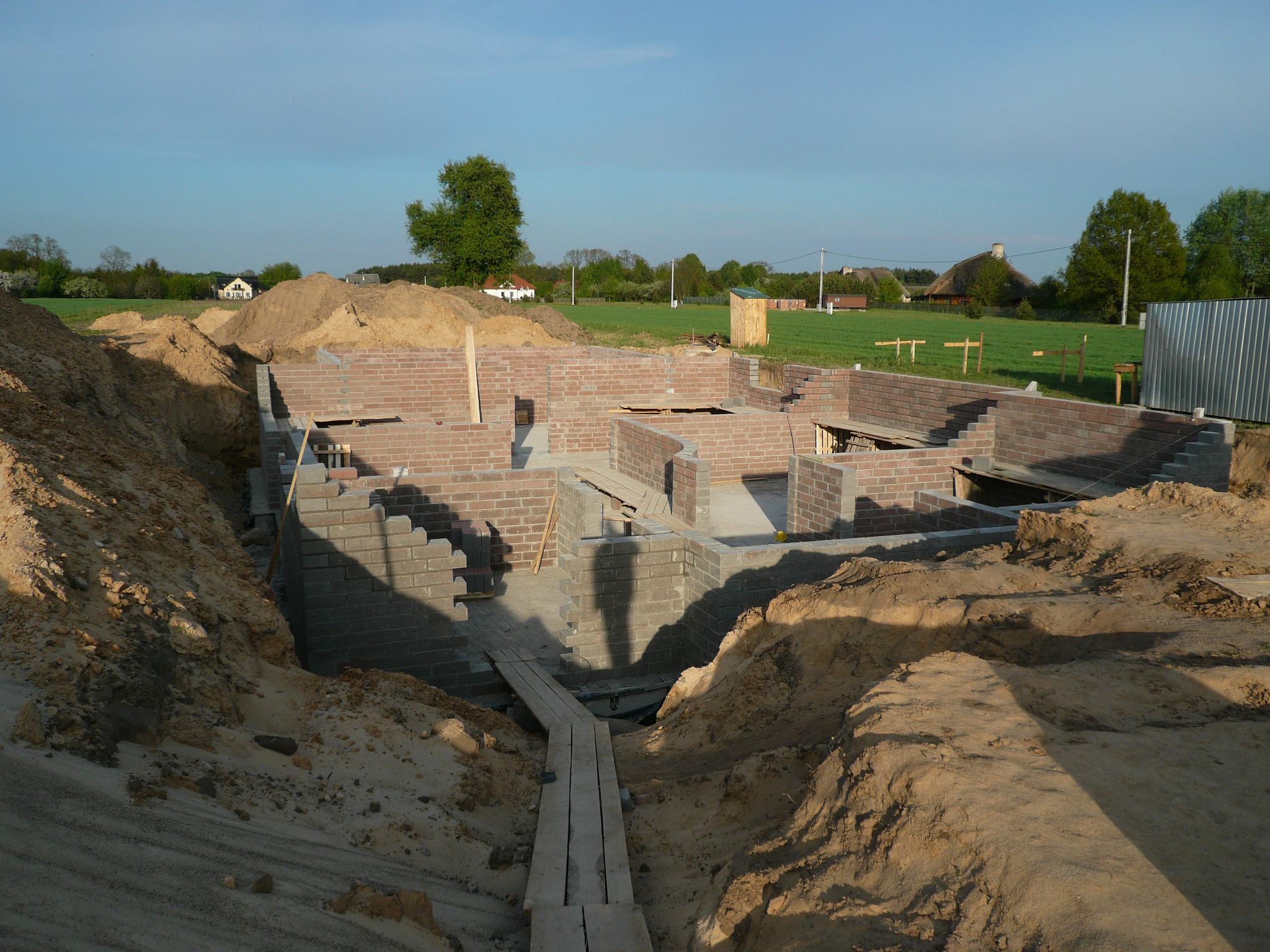 Wykop pod fundamenty z częściowo wymurowanymi ścianami z bloczków, widoczne drewniane pomosty i sterta piasku, w tle krajobraz wiejski.