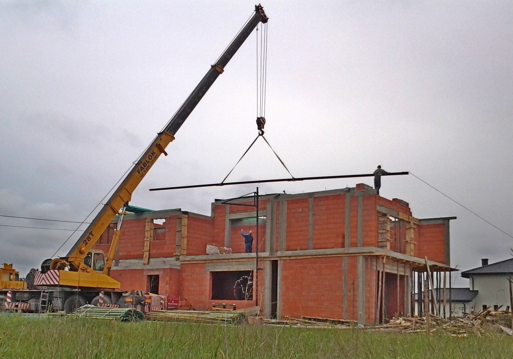 Budowa dwukondygnacyjnego domu murowanego z czerwonej cegły silikatowej, z użyciem żurawia wieżowego marki Fablok, dwóch robotników na budowie, jeden na dachu, drugi na rusztowaniu, widok z poziomu...