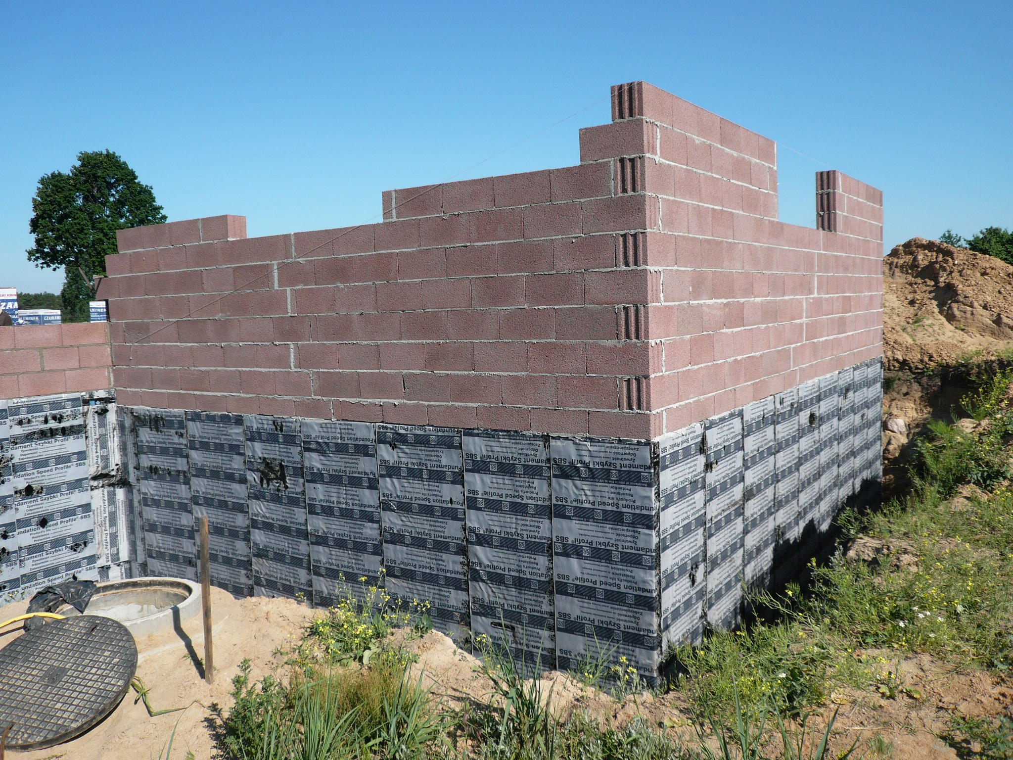 Surowe ściany parteru w trakcie budowy, wykonane z czerwonych bloczków, z widoczną hydroizolacją fundamentów, na tle zieleni i błękitnego nieba.