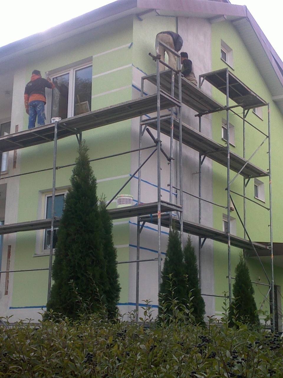 Pracownicy na rusztowaniu malują elewację domu na jasnozielono, z widocznymi białymi pasami i taśmą malarską zabezpieczającą okna, otoczenie budynku z niskimi iglakami.