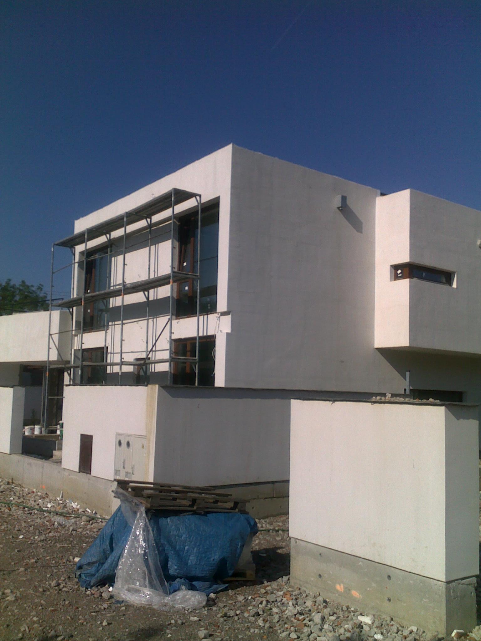 Dwupiętrowy dom murowany w trakcie ocieplania elewacji, widoczne rusztowania i materiały budowlane na ziemi.