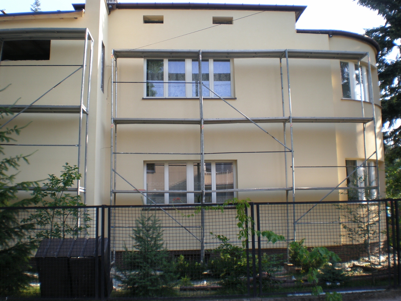 Jasna elewacja domu w trakcie prac, widoczne rusztowania i świeżo otynkowana ściana z oknami.