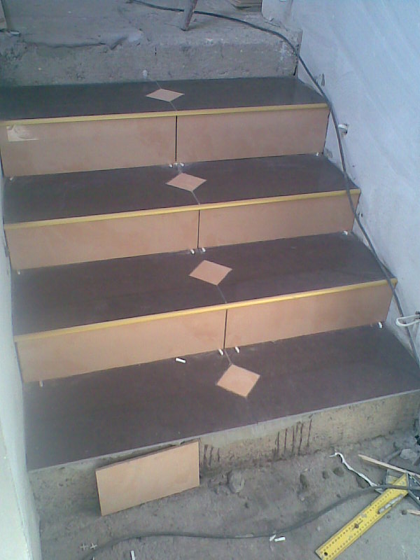 Układanie ciemnych płytek z rombowymi wstawkami na schodach wewnętrznych; widoczne narzędzia i materiały budowlane w trakcie prac.