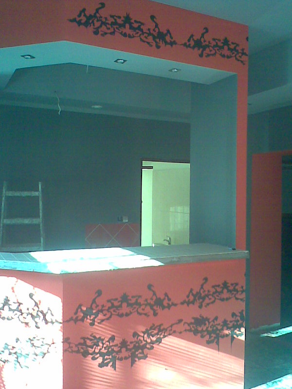 Wykończony bar z pomarańczowymi ścianami ozdobionymi czarnymi, abstrakcyjnymi wzorami, widoczny fragment sufitu z oświetleniem punktowym, w tle drabina i wejście do innego pomieszczenia.