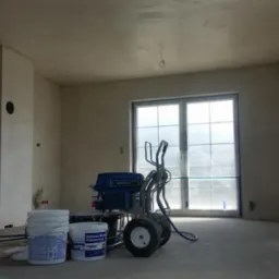 Wnętrze w trakcie prac wykończeniowych: ściany po szpachlowaniu, agregat malarski Graco i wiadra z gładzią polimerową. Okno zabezpieczone folią malarską.