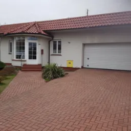 Dom jednorodzinny z czerwoną dachówką i podjazdem z kostki, widoczna elewacja w jasnym odcieniu, białe okna i drzwi wejściowe z numerem 19, garaż z bramą segmentową.