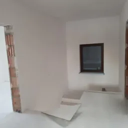 Surowe wnętrze z oknem i przygotowanymi otworami drzwiowymi, ściany po wstępnym malowaniu, podłoga zabezpieczona przed dalszymi pracami.