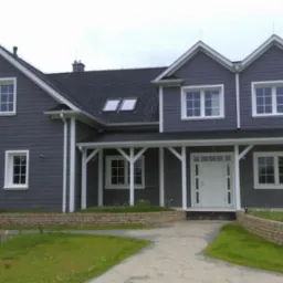 Dwukondygnacyjny dom z szarą elewacją, białymi oknami i kolumnami werandy, widoczny z przodu, z ciemnym dachem i dwoma oknami dachowymi.