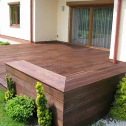 Nowoczesny taras drewniany z ciemnego drewna kompozytowego, z niską zabudową obok trawnika i domu. Widoczne detale desek i roślinność.