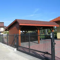 Drewniana wiata z dachem krytym gontem bitumicznym, widoczna zza czarnego, metalowego ogrodzenia posesji w słoneczny dzień. Na drugim planie budynki mieszkalne.