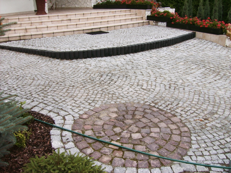 Granitowa kostka brukowa ułożona w okrągły wzór z ciemniejszego kamienia, obrzeża z drewnianych palisad, schody wejściowe do domu w tle, widoczny fragment węża ogrodowego.
