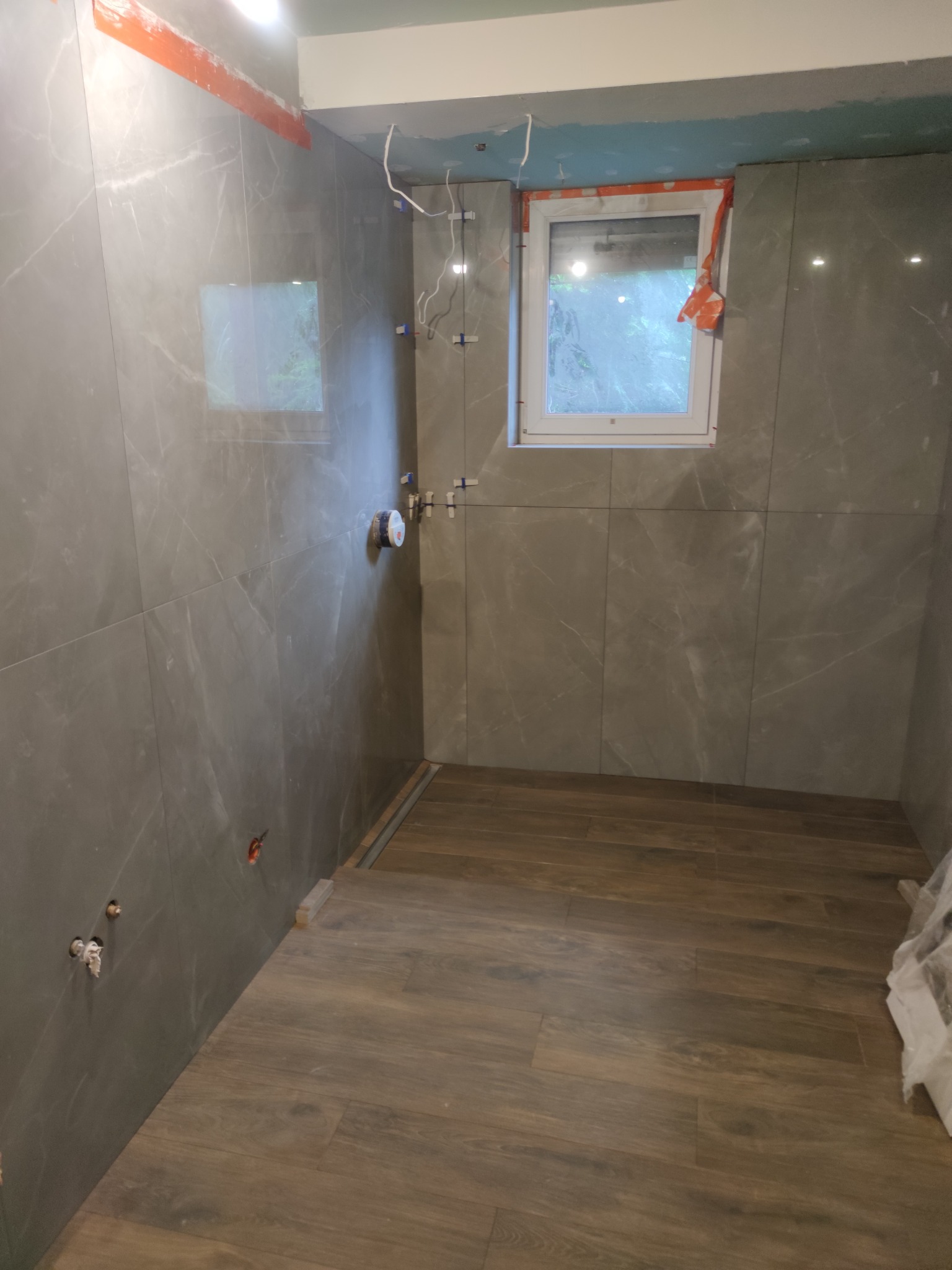 Wnętrze łazienki w trakcie wykańczania: szare płytki imitujące marmur na ścianach, drewnopodobna podłoga, okno z widokiem na zieleń, widoczne instalacje i elementy montażowe.