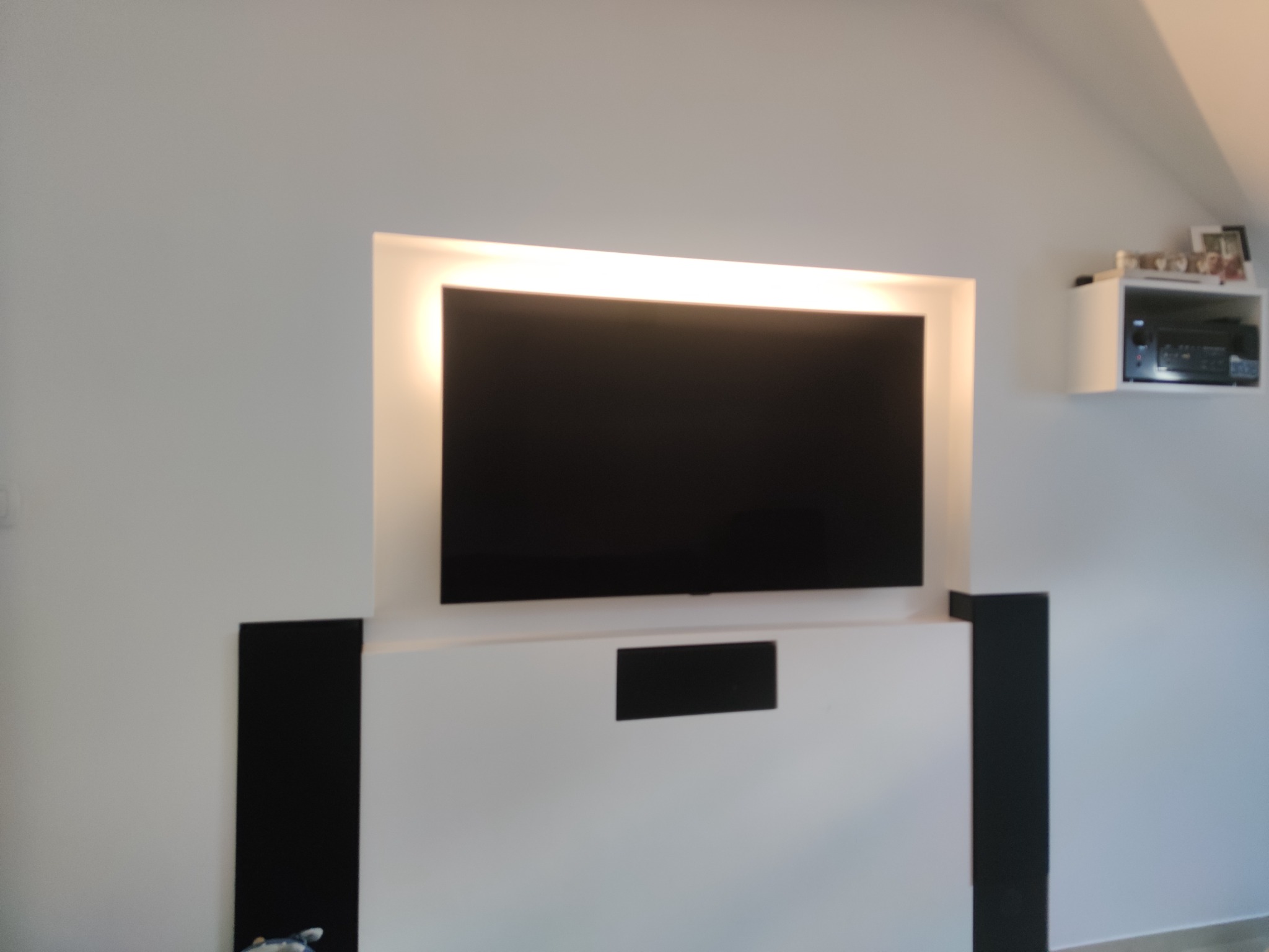Ściana z wnęką pod telewizor, podświetlona od góry, z czarnymi elementami dekoracyjnymi po bokach i półką na sprzęt RTV w górnym rogu.
