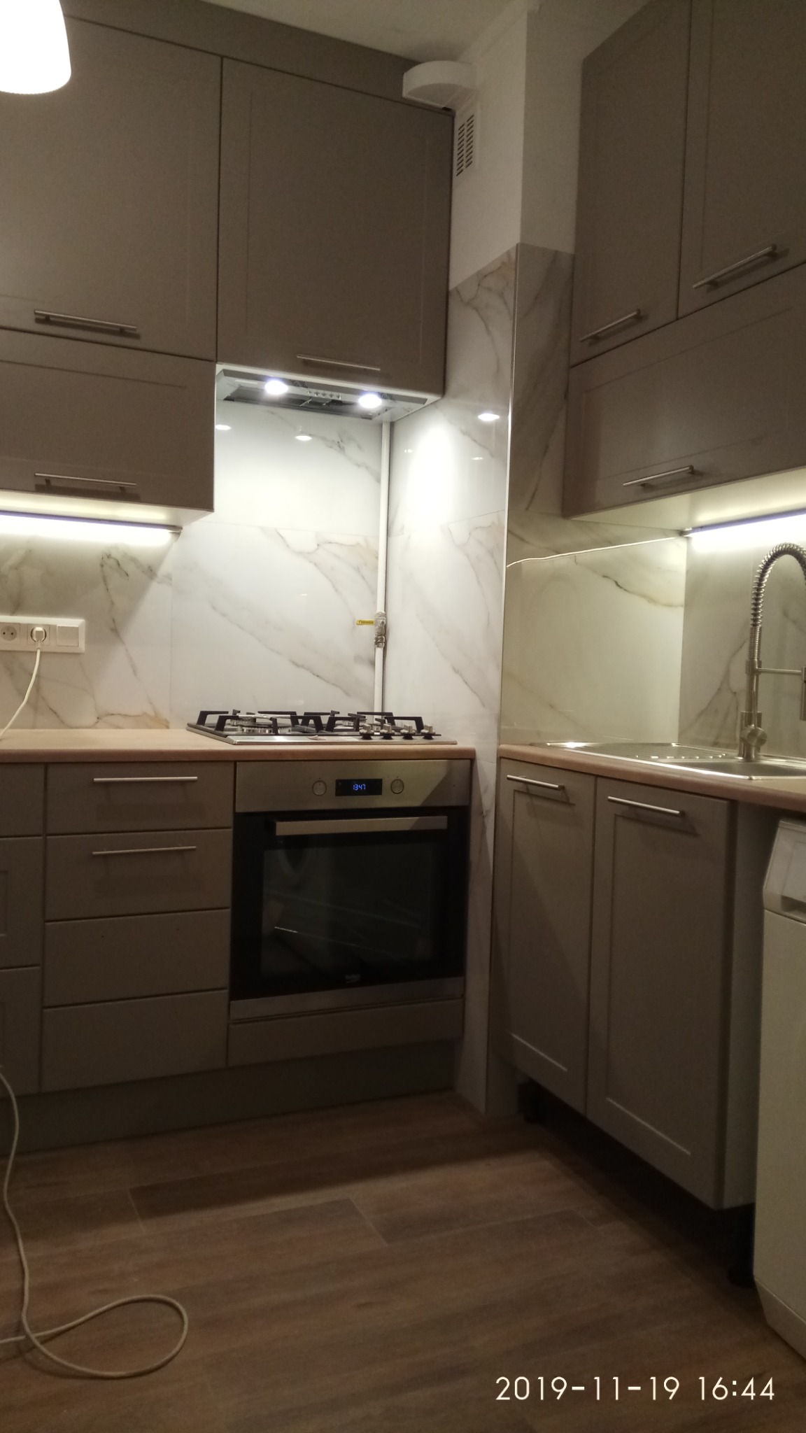 Szara zabudowa kuchenna z marmurowymi płytkami, oświetleniem LED pod szafkami i piekarnikiem z płytą gazową.