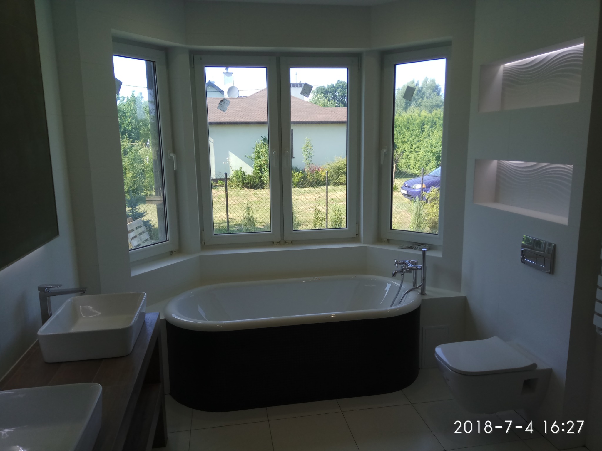 Nowoczesna łazienka z wolnostojącą wanną na tle okna z widokiem na ogród, minimalistyczna umywalka nablatowa i podwieszana toaleta.