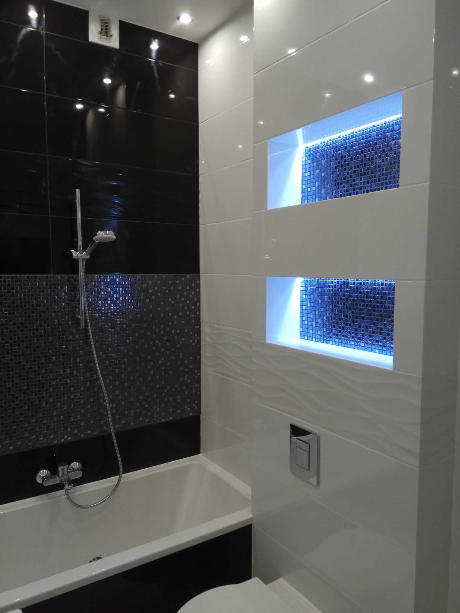 Nowoczesna łazienka z czarno-białymi płytkami, podświetlanymi wnękami z mozaiką oraz armaturą.