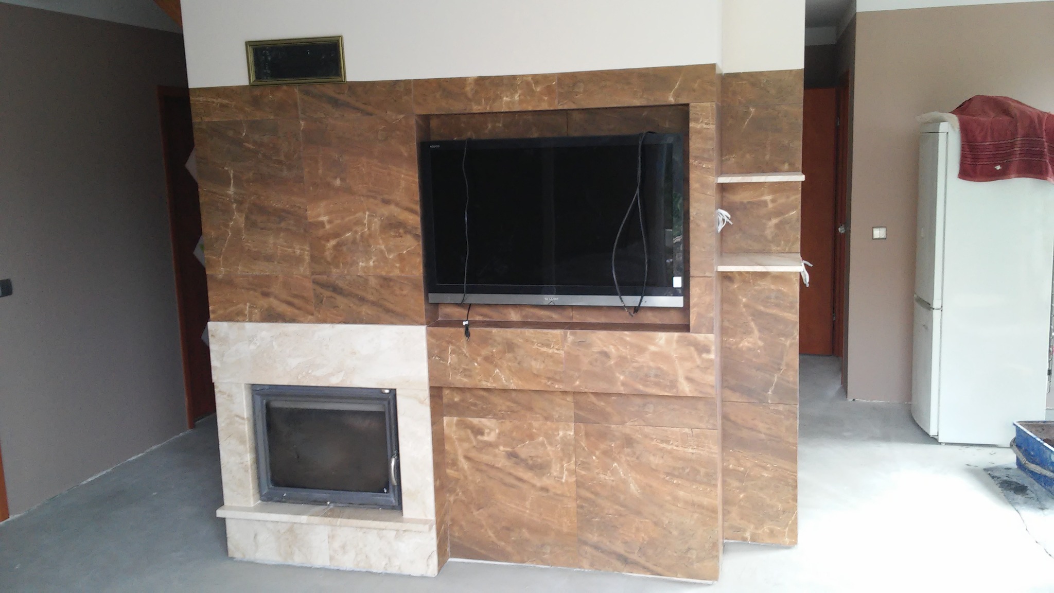 Obudowa kominka z marmurową okładziną, wnęka na telewizor z widocznymi kablami, półki z marmuru po prawej stronie, całość w surowym stanie, podłoga nie wykończona.