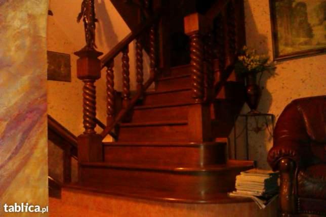 Drewniane schody z toczonymi balustradami w ciepłym oświetleniu. Widoczny fragment ściany z wzorem i skórzany fotel.