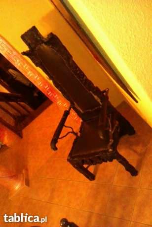 Ciemne, bogato zdobione krzesło z tapicerowanym siedziskiem, ustawione ukośnie na podłodze. Widoczny fragment innego mebla w tle. Oświetlenie górne.