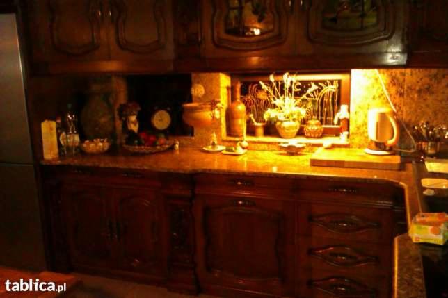 Ciemne, lakierowane fronty szafek kuchennych z frezowanymi zdobieniami i kamienny blat. Na blacie dekoracje i akcesoria kuchenne. Styl tradycyjny, ciepłe oświetlenie.