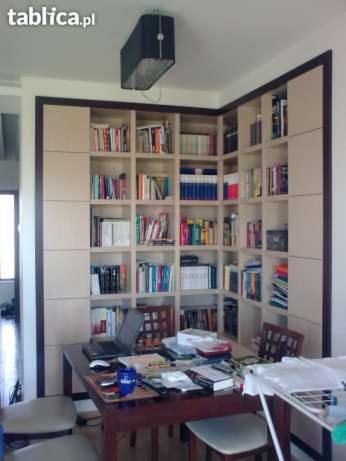 Jasna, narożna biblioteczka z ciemnym obramowaniem, wypełniona książkami, nad biurkiem z laptopem. Widok na wnętrze pokoju, z lampą sufitową.