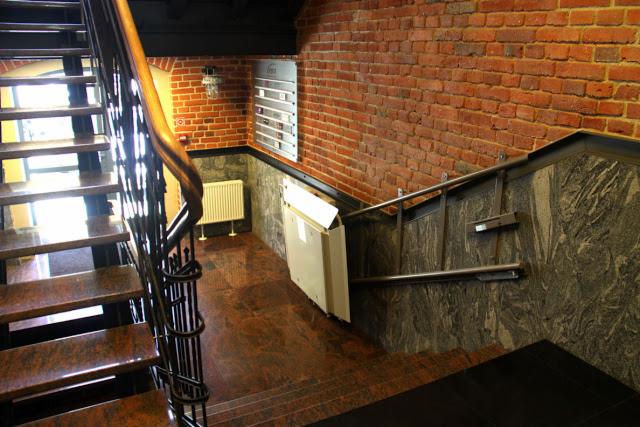 Klatka schodowa z metalową balustradą i schodami z czerwonego granitu. Ściany z czerwonej cegły i marmuru. W tle grzejnik i tablica informacyjna.