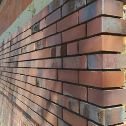 Ceglana ściana zewnętrzna w trakcie murowania, widoczne termoizolacja, poziomica i zbiornik na wodę w tle.