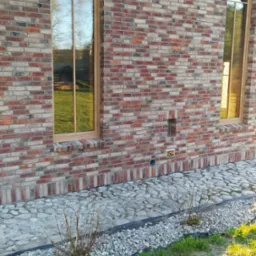 Fragment elewacji budynku w Nowej Wsi Lęborskiej z cegły klinkierowej w odcieniach czerwieni i szarości, z dwoma oknami o nietypowym kształcie i brązowych ramach. U podnóża ściany widoczny kamienny...