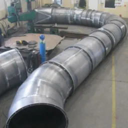 Długi, spawany, metalowy rurociąg w kształcie litery S, z widocznymi spawami i obręczami, leżący na betonowej podłodze hali produkcyjnej.