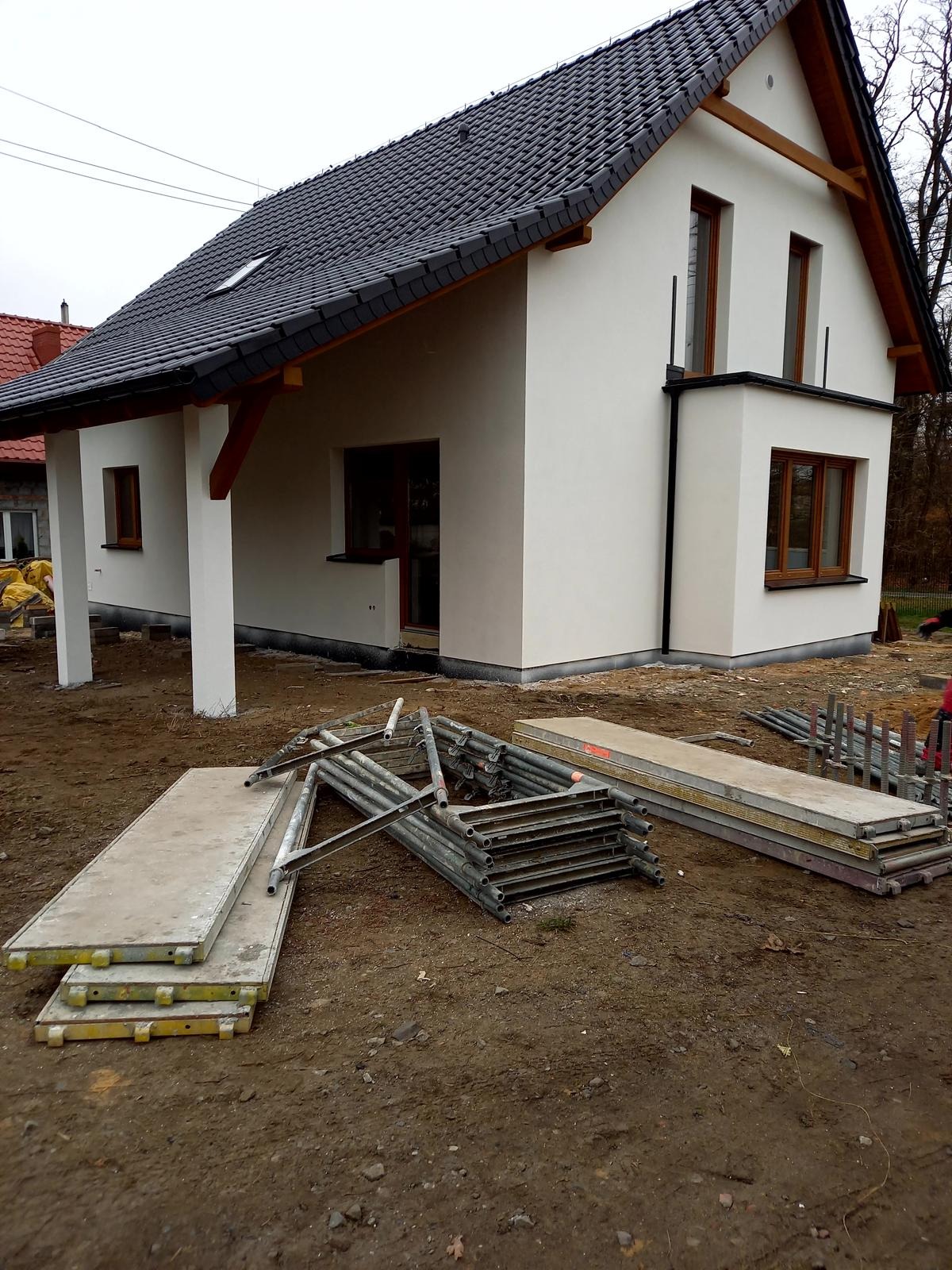 Nowo wybudowany, parterowy dom z białym tynkiem i ciemną dachówką, widoczne rusztowania i materiały budowlane na ziemi, okna z brązowymi ramami, brak zagospodarowania terenu.