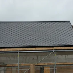 Fragment dachu krytego ciemną dachówką rombową, widoczne rusztowanie i surowy mur budynku w trakcie budowy.