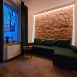 Salon z ceglaną ścianą, oświetleniem LED i zieloną sofą. Parkiet na podłodze, zasłony w oknie. Minimalistyczny stolik kawowy. Ciepłe, stonowane oświetlenie.