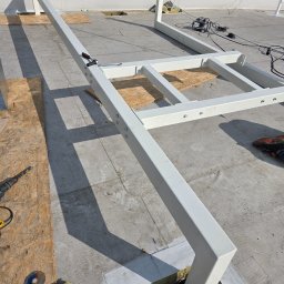 PIT-BUD Piotr Kudliński - Biała, metalowa konstrukcja na płaskim dachu z widocznymi narzędziami i elementami montażowymi. W tle zielony krajobraz.