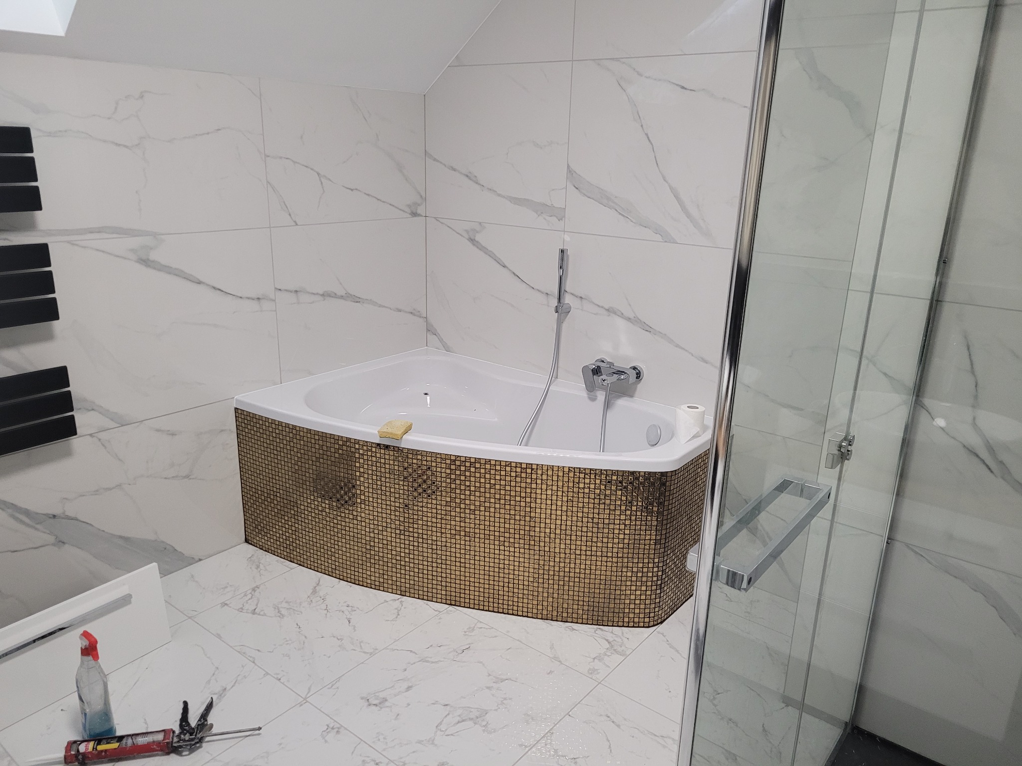 Nowoczesna łazienka z wanną narożną obłożoną mozaiką w odcieniach złota, białe marmurowe płytki na ścianach i podłodze, kabina prysznicowa ze szklanymi drzwiami oraz akcesoria i narzędzia...