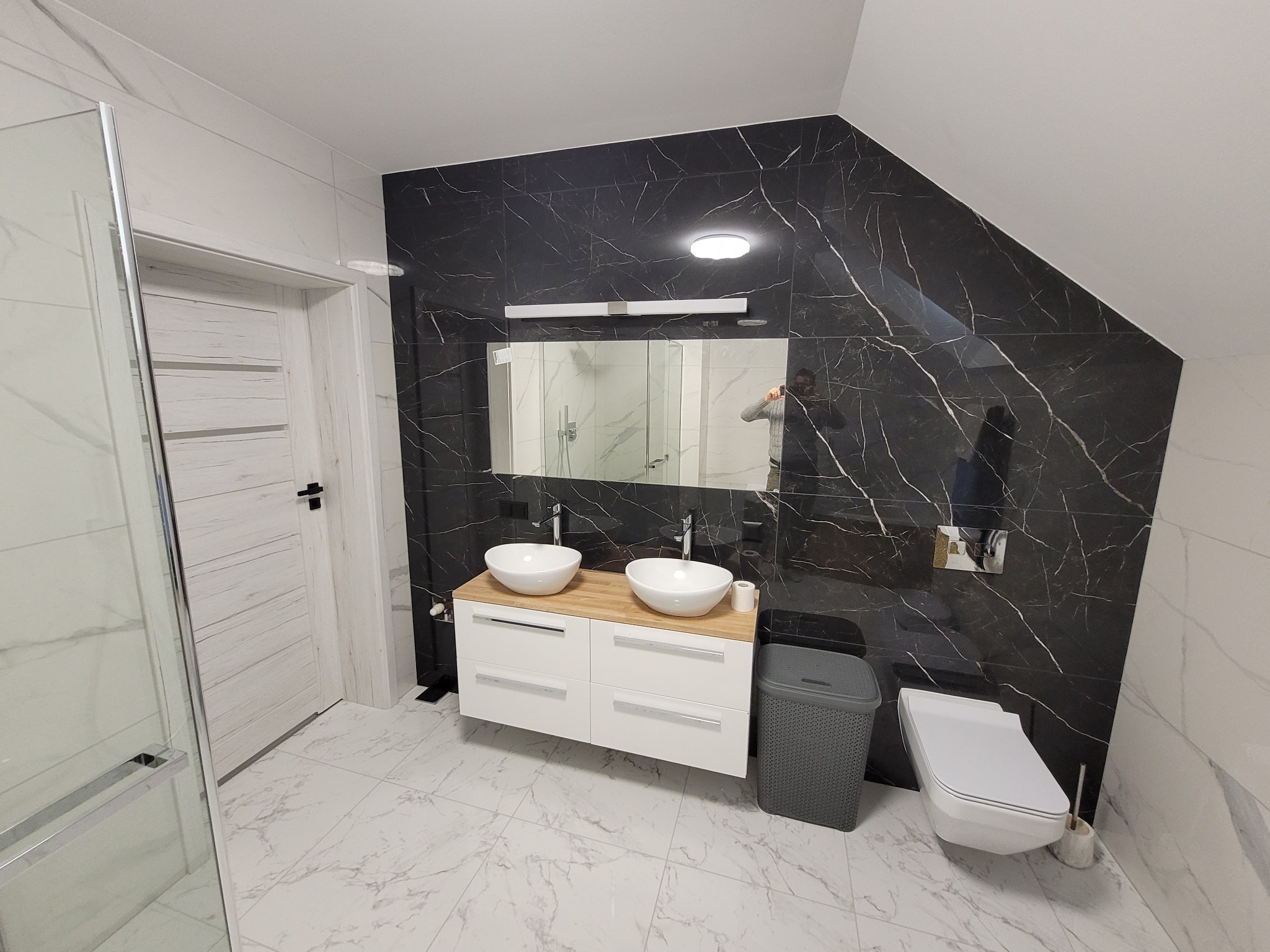 Nowoczesna łazienka na poddaszu z czarno-białymi płytkami imitującymi marmur, podwójną umywalką nablatową na szafce z drewnianym blatem i wiszącą toaletą.