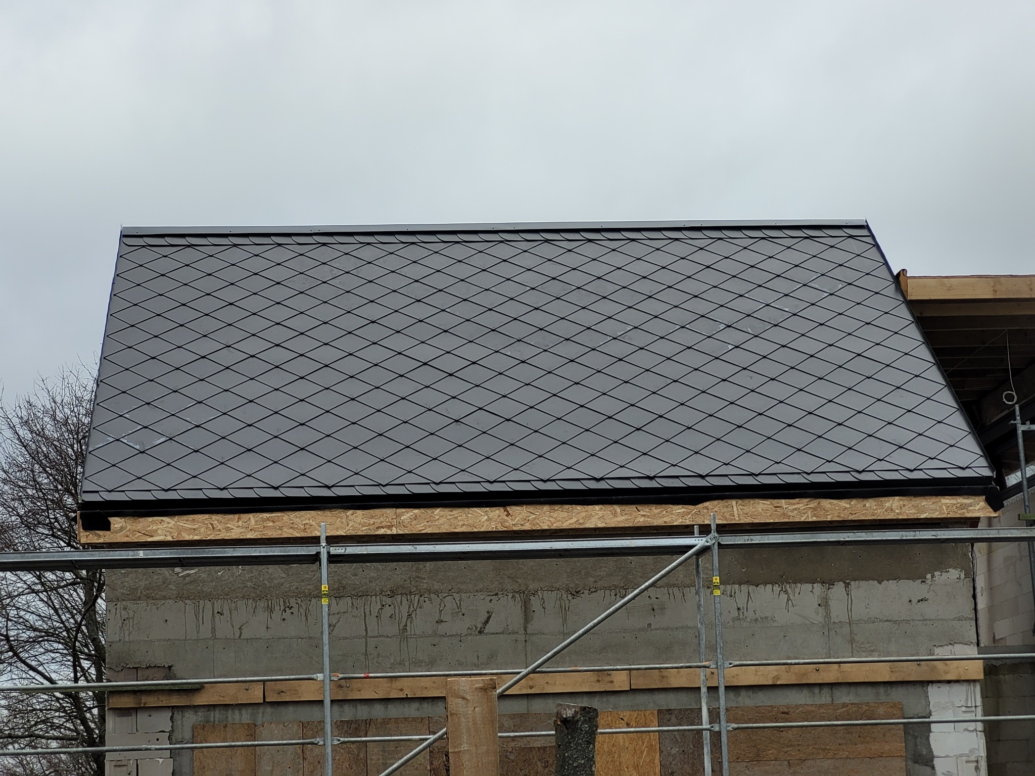 Fragment dachu krytego ciemną dachówką rombową, widoczne rusztowanie i surowy mur budynku w trakcie budowy.