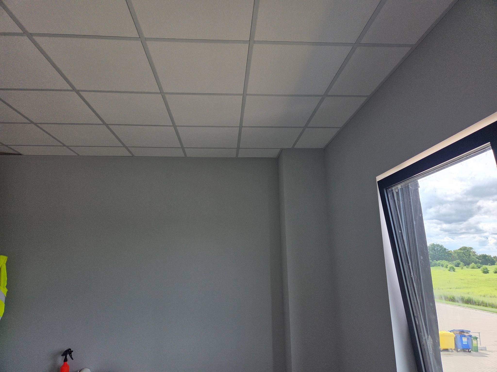 Szara ściana z oknem i zamontowanym podwieszanym sufitem kasetonowym. Widoczne narzędzia i fragment odblaskowej kamizelki. Widok na zewnątrz przez okno.
