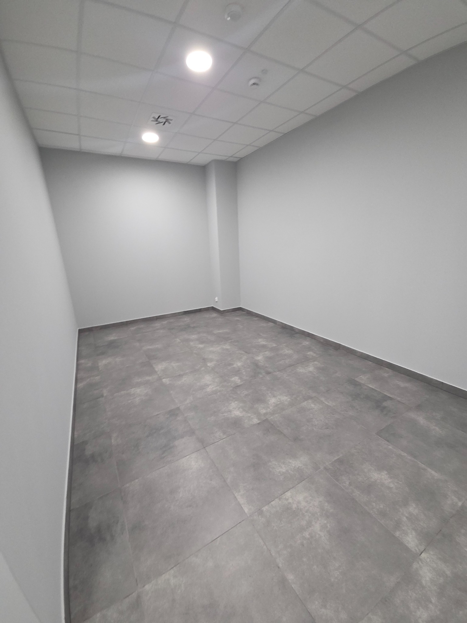 Szare, prostokątne płytki podłogowe w pustym, pomalowanym na szaro pomieszczeniu z sufitową kratownicą i oświetleniem LED. Perspektywa z narożnika.