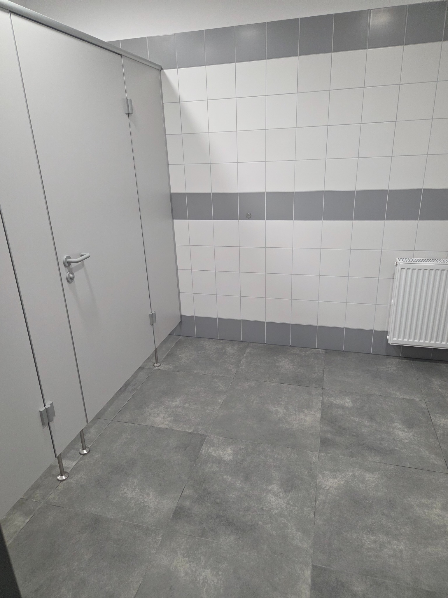 Wykończona toaleta publiczna z szarymi płytkami podłogowymi i białymi płytkami ściennymi z szarymi pasami. Widoczne kabiny WC i grzejnik.